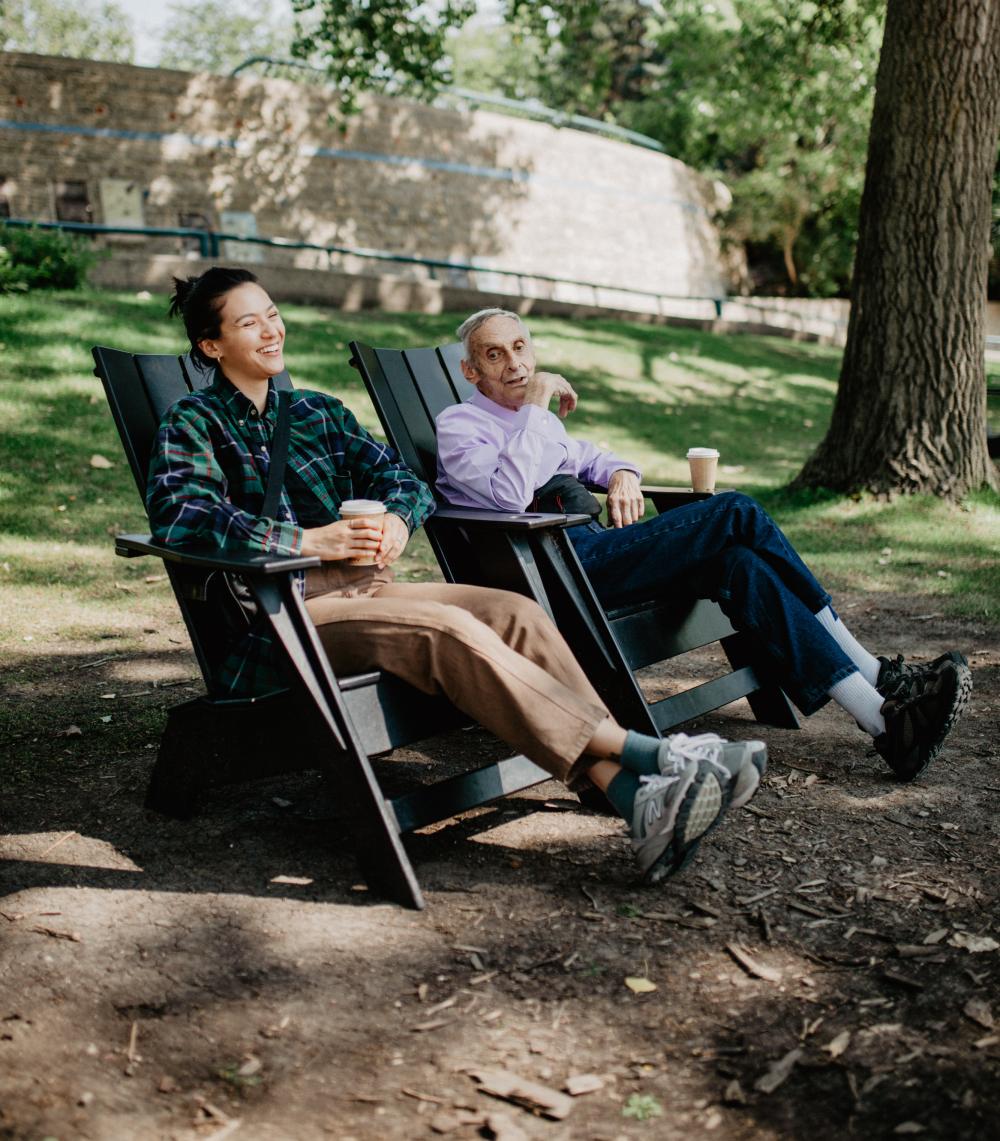 two people sitting on wooden chairs drinking coffee outside